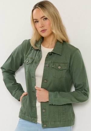 Blonde vrouw met een groene jas met knopen over een beige overhemd, staand met één hand in de zak tegen een witte achtergrond.