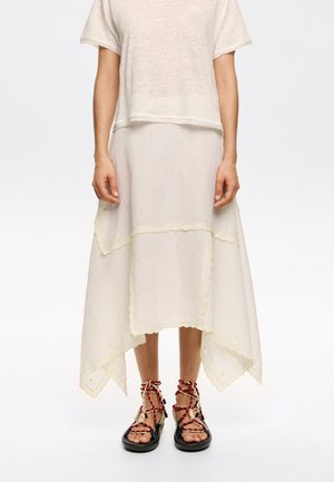 Kleid aus Leinenmischung in Creme, mit gewelltem Saum, gelben Blumenakzenten und einem mehrlagigen Rock. Kombiniert mit schwarzen Sandalen und roten Riemen.