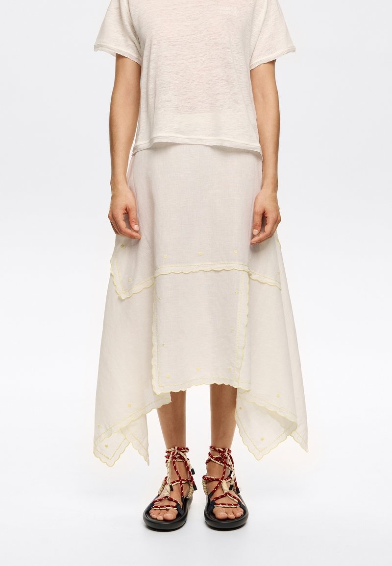 Linen blend dress in cream, featuring scalloped hem, yellow floral accents, and a layered skirt. Paired with black sandals and red straps.