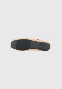 Platt beige toffel med svart gummisula, mjuk fluffig textur och spännedetalj som visas från undersidan på en vit bakgrund.