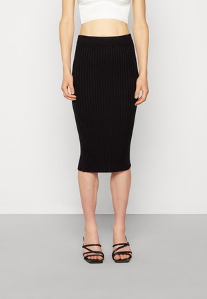 Femme portant une jupe midi côtelée noire, un top court blanc et des sandales noires à brides ouvertes, debout sur un sol gris contre un mur blanc.