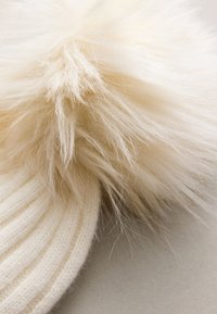 Cappello a cuffia bianco lavorato a maglia con un grande pompon in pelliccia sintetica soffice sulla cima, che mostra una texture morbida e sottili variazioni di colore.