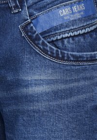 Tmavě modré džíny s hladkou texturou, s postranní kapsou a štítkem s nápisem "CARS JEANS" poblíž pásku.