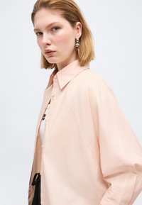 Νεαρή γυναίκα με κοντά ξανθά μαλλιά φοράει ανοιχτό ροζ πουκάμισο με κουμπιά και μεγάλα ασημένια σκουλαρίκια, κοιτάζοντας αριστερά με ουδέτερη έκφραση.