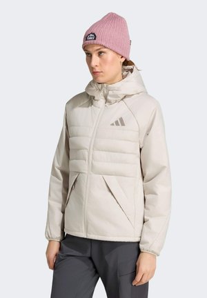 Frau trägt eine hellbeige gesteppte Jacke mit Kapuze und dunkle Hose, ergänzt durch eine rosa gestrickte Mütze.