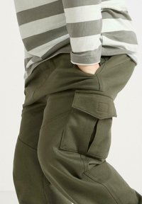 Olijfgroene cargobroek van stof met een zijzak en een gladde textuur. Gepaard met een grijs-witte gestreepte lange mouw shirt.