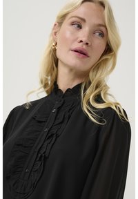 Črna bluzica z nabranim sprednjim delom, visokim ovratnikom in gumbom za zapiranje. Narejena iz lahkega blaga z prosojnimi poudarki na rokavih.