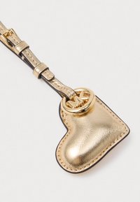 Charm in pelle oro a forma di cuore con tracolla regolabile e anello metallico con logo MK dorato su uno sfondo chiaro.