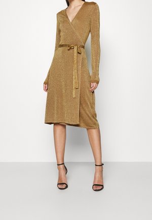 Vrouw draagt een knielange, lange mouw, metallic gouden wikkeljurk met een gestrikte riem en zwarte open teen hoge hakken.