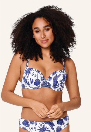 Kvinne med krøllete hår iført en hvit og blå bikini med blomsterprint, stående med hendene løst sammenlåst foran mot en ensfarget bakgrunn.