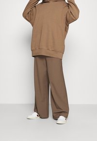 Felpa marrone oversize con tasca frontale, abbinata a pantaloni marroni a gamba larga. Sneakers bianche con dettagli rossi completano il look.