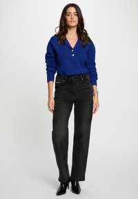 Suéter azul de punto con escote en V, adornado con un botón en forma de corazón, combinado con jeans negros de cintura alta y botas negras de punta.