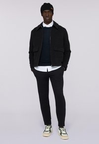 Giacca zip grigia scura con due tasche frontali, indossata sopra un maglione blu navy e una camicia bianca con colletto. Abbinata a pantaloni neri a sigaretta e sneakers.