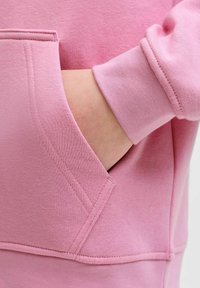 Lyserød sweatshirt med en diagonal lomme med synlige sømme og blødt stof, der fremviser en glat tekstur og et afslappet design.