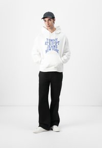 Sudadera blanca con texto azul que dice "TOMMY ATH.DIPT JEANS NEW YORK," combinada con pantalones negros y zapatillas blancas, de pie sobre un fondo blanco.