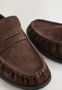 Bruine suède loafers met een afgeronde neus, moccasin-stiksel en geplooide accenten langs de kraag. Zwarte rubberen zool. Gestructureerde afwerking.