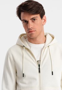 Witte hoodie met ritssluiting en een capuchon met trekkoord, voorzien van een zwarte rits en tips. Draagt over een lichtgekleurde shirt met een grafische print.