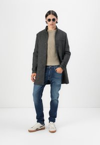 Manteau en mélange de laine gris avec un devant à boutons, pull en tricot beige, jean slim bleu et chaussures de sport blanches avec des accents marron. Lunettes de soleil rondes.