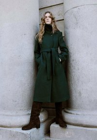 Mujer con cabello largo y ondulado apoyada entre grandes columnas de piedra, vistiendo un abrigo verde oscuro con cinturón y botas altas de color marrón oscuro.