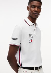 Άνδρας που φοράει λευκό πόλο μπλουζάκι Tommy Hilfiger με λογότυπα Cadillac και Pirelli, κόκκινες και μπλε ρίγες στα πλάγια και στο γιακά, στέκεται μπροστά σε λευκό φόντο.