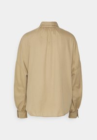 Blouse beige clair avec un col classique, un détail froncé au dos et des manches bouffantes se terminant par des poignets ajustés. Tissu lisse et texturé.