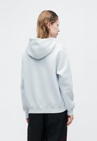 Jasnoniebieska bluza z kapturem z miękkiego materiału, o luźnym kroju, z kapturem na sznurki oraz ściągaczami na rękawach i dolnym brzegu. Noszona z ciemnymi spodniami.