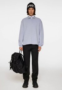 Polo a maniche lunghe grigio chiaro con colletto, abbinata a pantaloni neri e uno zaino nero grande. Berretto nero con scritta bianca.