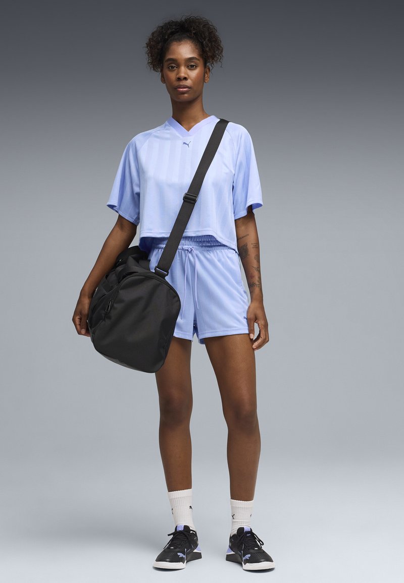 Γυναίκα που φοράει αθλητικά ρούχα ανοιχτού μωβ και μαύρα αθλητικά παπούτσια, κρατώντας μια μαύρη τσάντα γυμναστηρίου στον ώμο της, στέκεται μπροστά σε γκρι φόντο.