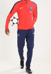 Punainen vetoketjullinen takki sinisillä yksityiskohdilla ja Pariisi-logo, yhdistettynä tummansinisiin housuihin. Musta jalkapallo pidetään vasemmassa kädessä, mustat ja valkoiset pelikengät.