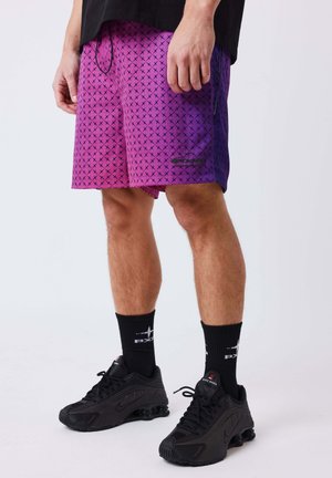 Mannelijke benen met paarse korte broek met patroon, zwarte sportsokken met witte tekst en zwarte Nike Shox sneakers op een effen achtergrond.