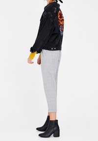 Veste en denim noire avec broderies colorées au dos, assortie avec un pantalon à carreaux gris et des bottines noires. Textures lisses et coupe ajustée.