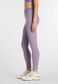 Femme portant des leggings taille haute lilas et des baskets blanches, debout de profil sur un fond clair uni.