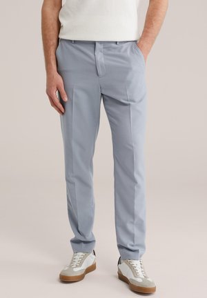 Un homme portant un pantalon bleu clair sur mesure, des baskets blanches avec des semelles beige et marron, et un haut sans manches blanc texturé, debout les mains dans les poches.