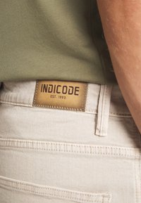 Bež hlače s kožnom etiketom na kojoj piše "INDICODE EST. 1993." Tkanina je glatka s vidljivim detaljima šivanja i uskim pojasom.