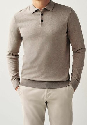 Mann trägt taupefarbenen Langarm-Polo-Pullover mit schwarzen Knöpfen, Hände in den Taschen, kombiniert mit hellbeigen Hosen vor einfachem Hintergrund.