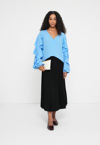 Blouse bleu clair avec des manches à volants, décolleté en V et fermeture à boutons, associée à une jupe plissée noire et une pochette crème.