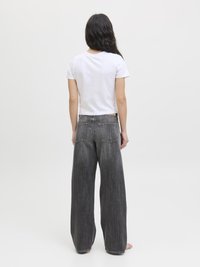 Kvinne med langt mørkt hår iført en hvit kortermet topp og løstsittende falmede sorte jeans, stående barføtt mot en hvit bakgrunn.