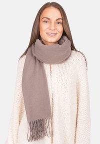 Femme souriante avec des cheveux bruns lisses portant un pull en tricot beige et une épaisse écharpe taupe avec des franges d'un côté.