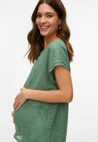 Top de encaje verde con mangas cortas, que presenta un patrón texturizado. La modelo sostiene un abdomen embarazado, mostrando un ajuste relajado.