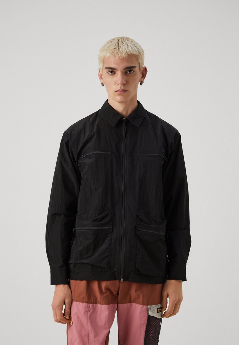 Joven con cabello corto rubio, vestido con una chaqueta negra con cremallera y pantalones cargo bicolor en rosa, marrón y negro, de pie frente a un fondo simple.