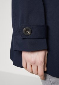 Rękaw granatowej kurtki z detalem guzika. Posiada ustrukturyzowany mankiet, gładką teksturę i subtelne przeszycia. Dłoń oparta na rękawie.