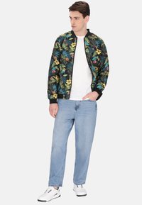 Jachetă bomber florală cu modele multicolore pe un fundal negru, asortată cu blugi bleu deschis și adidași albi.
