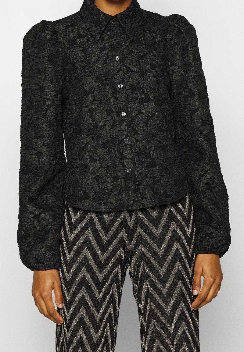 Blouse noire texturée à motifs floraux, boutonnée, avec manches longues bouffantes, associée à un pantalon noir et argent à motif chevrons.