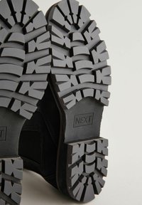 Las suelas de las botas negras cuentan con profundas ranuras para una buena tracción. El material de goma proporciona durabilidad y una superficie texturizada para un mejor agarre. La marca es visible.
