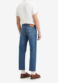 Jeans de mezclilla azul con corte de pierna recta, cintura alta y parche trasero de cuero. Combinados con una camiseta blanca y zapatos marrones.