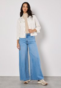Chaqueta de mezclilla blanca con bolsillos frontales, suéter de cuello alto beige y jeans de pierna ancha azul claro, combinados con zapatillas beige y marrones.