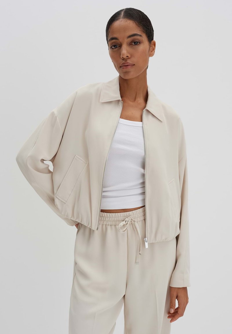 Beige kurze Jacke mit Kragen, Frontreißverschluss und Seitentaschen. Glatter Stoff mit entspannter Passform und weiten Ärmeln.