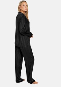Svart satin pyjamas-set med vertikala ränder, innehållande en långärmad topp och löst sittande byxor, accentuerat med vit piping.