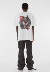 Biely grafický tričko s detailným dizajnom draka a červenými akcentmi, nosené s čiernymi cargo nohavicami. Voľný strih, krátke rukávy, bez viditeľných log. 
