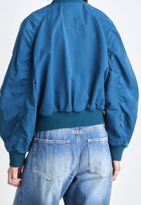 Blå bomberjacka med ribbad krage och mudd, med voluminösa ärmar och en rynkad nertill, tillsammans med ljusa jeans.